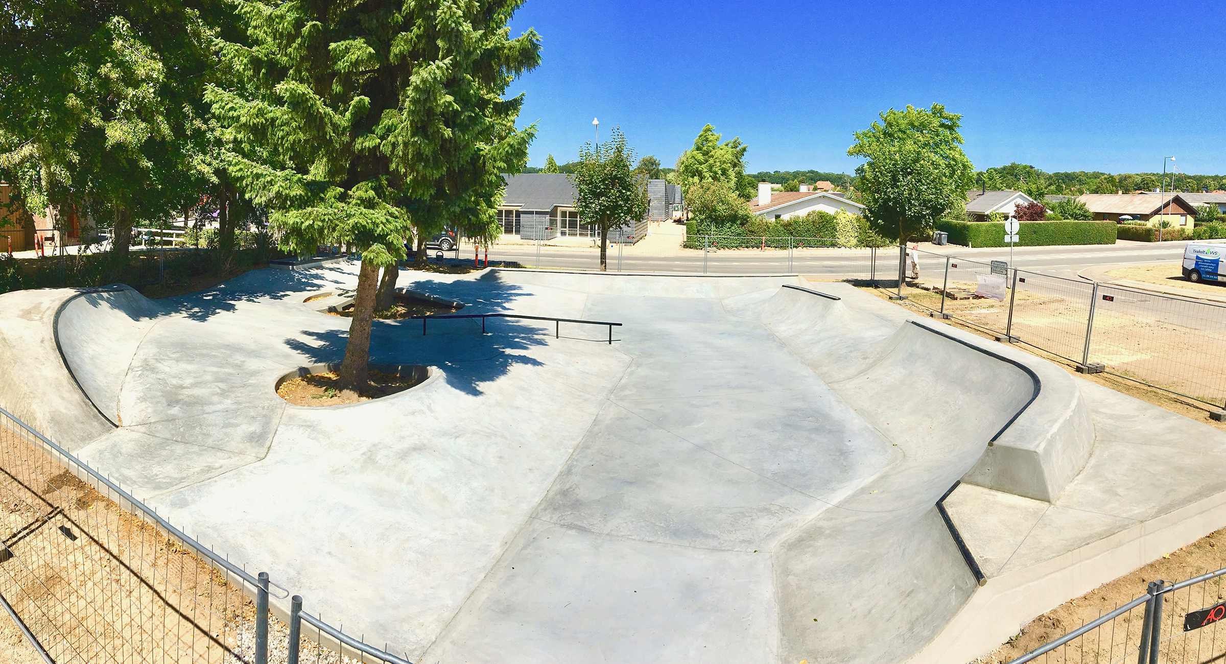 Billedet viser en indhegnet skatepark med forskellige buede betonramper og et rail