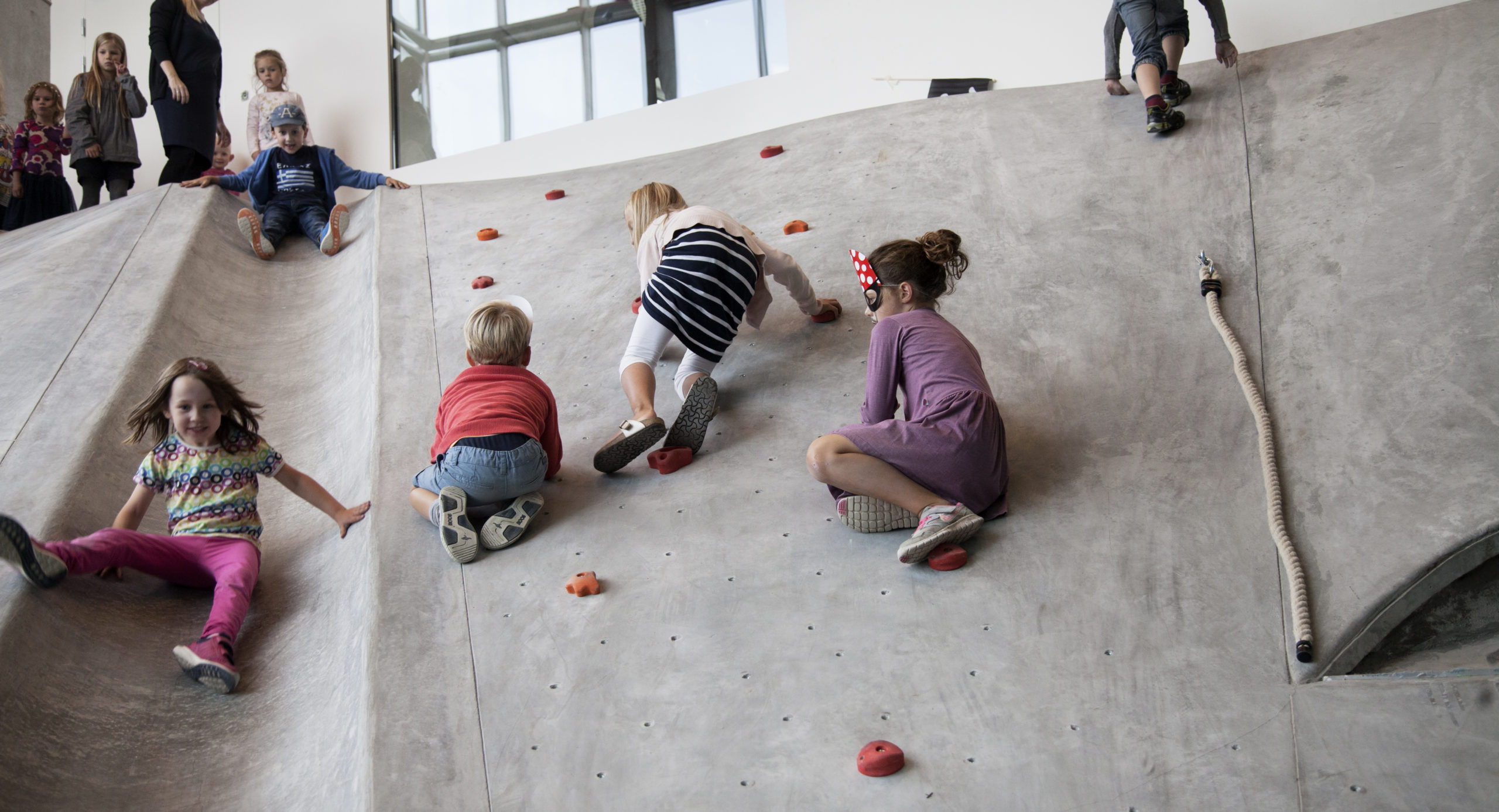 Børn klatrer op ad en skrå betonvæg ved hjælp af indbyggede klatregreb. Ved siden af rutscher børn ned ad en støbt rutschebane i betonoverfladen
