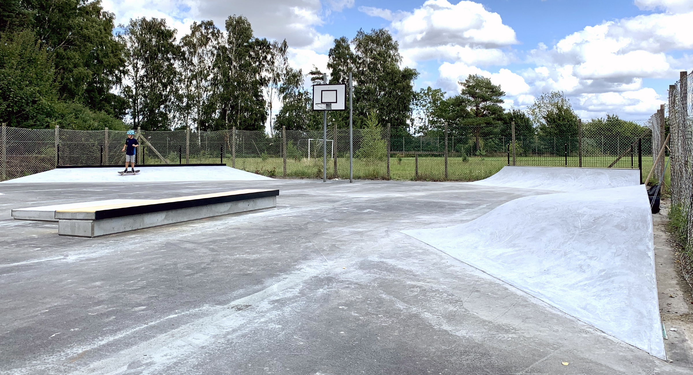I forgrunden ses et hip og en curb i beton med en manual pad bagved. I baggrunden ses en basketkurv og to hjørneramper i beton