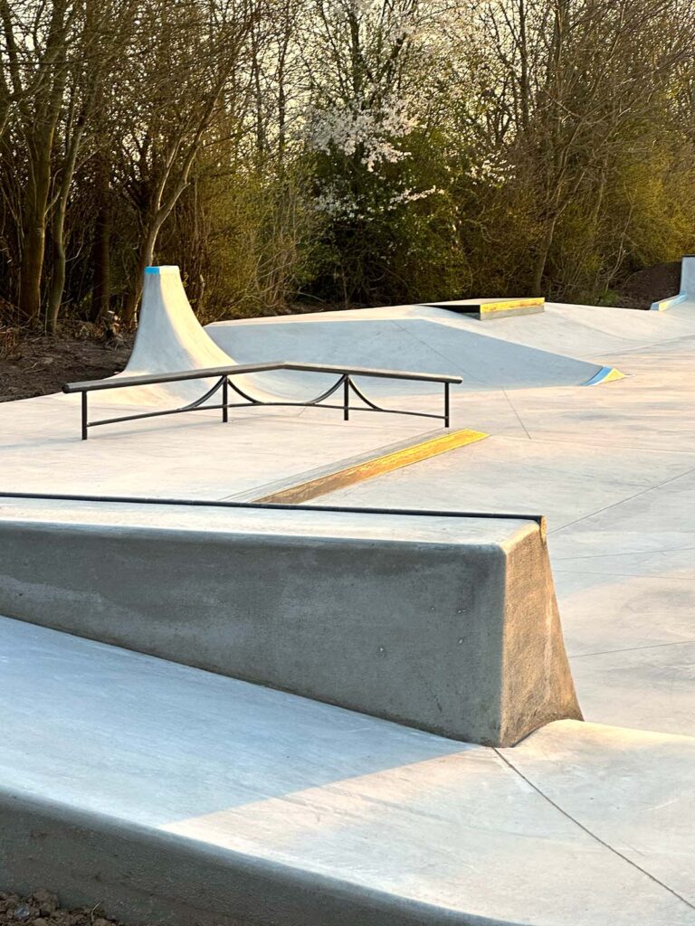 Foto på tværs af Middelfart Skatepark, hvor man både ser bagsiden af en quarterpipe, et sort rail, en curb på toppen af en bank og en høj, smal quarterpipe
